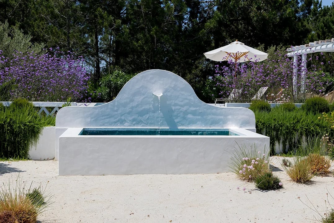 Outdoor white water fountain with greenery and purple flowers at Rafter Comporta Celeste in Comporta, Portugal