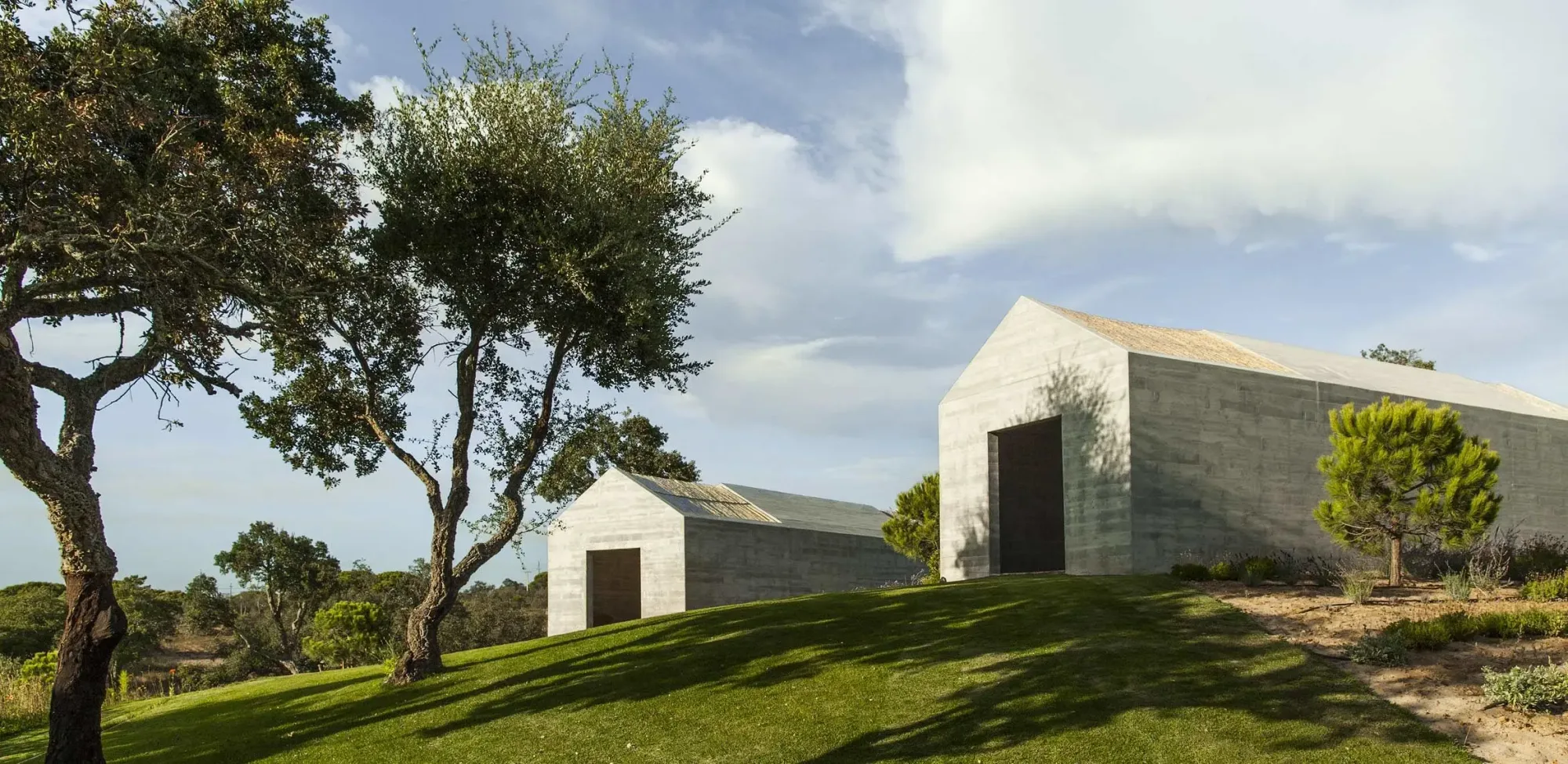Two minimalist concrete cabins on a grassy hillside with trees at Rafter Melides Atlantica in Melides, Portugal